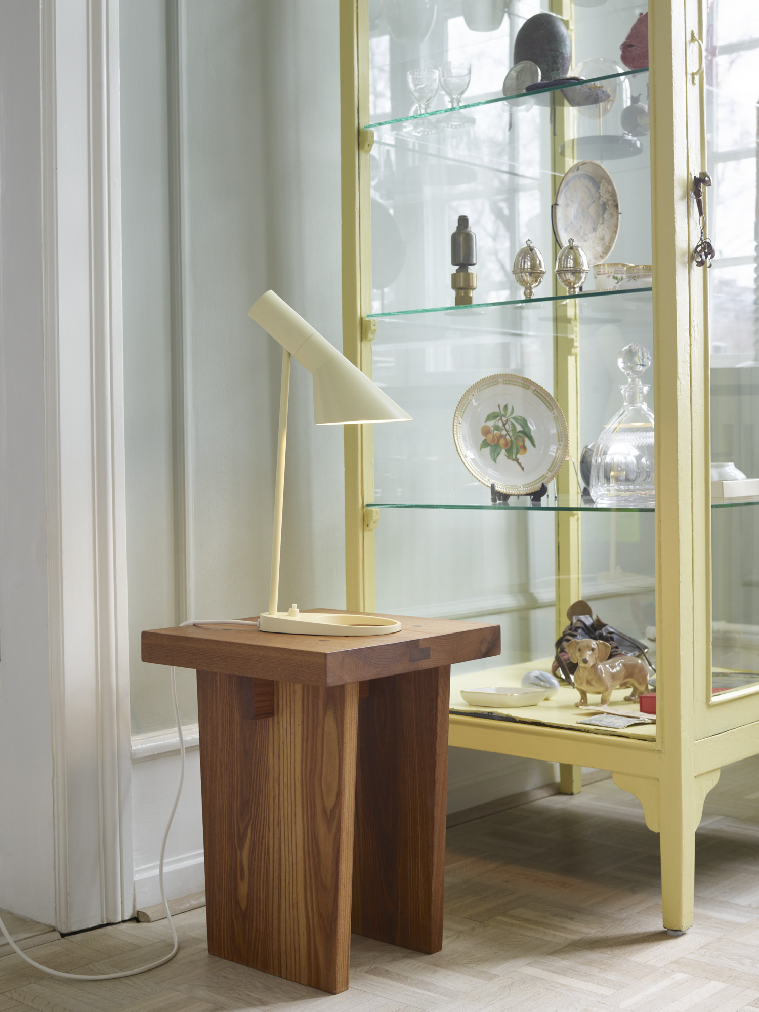 AJ Mini Table lamp in soft lemon on a wooden table beside a yellow display cabinet, © 2025 Louis Poulsen.