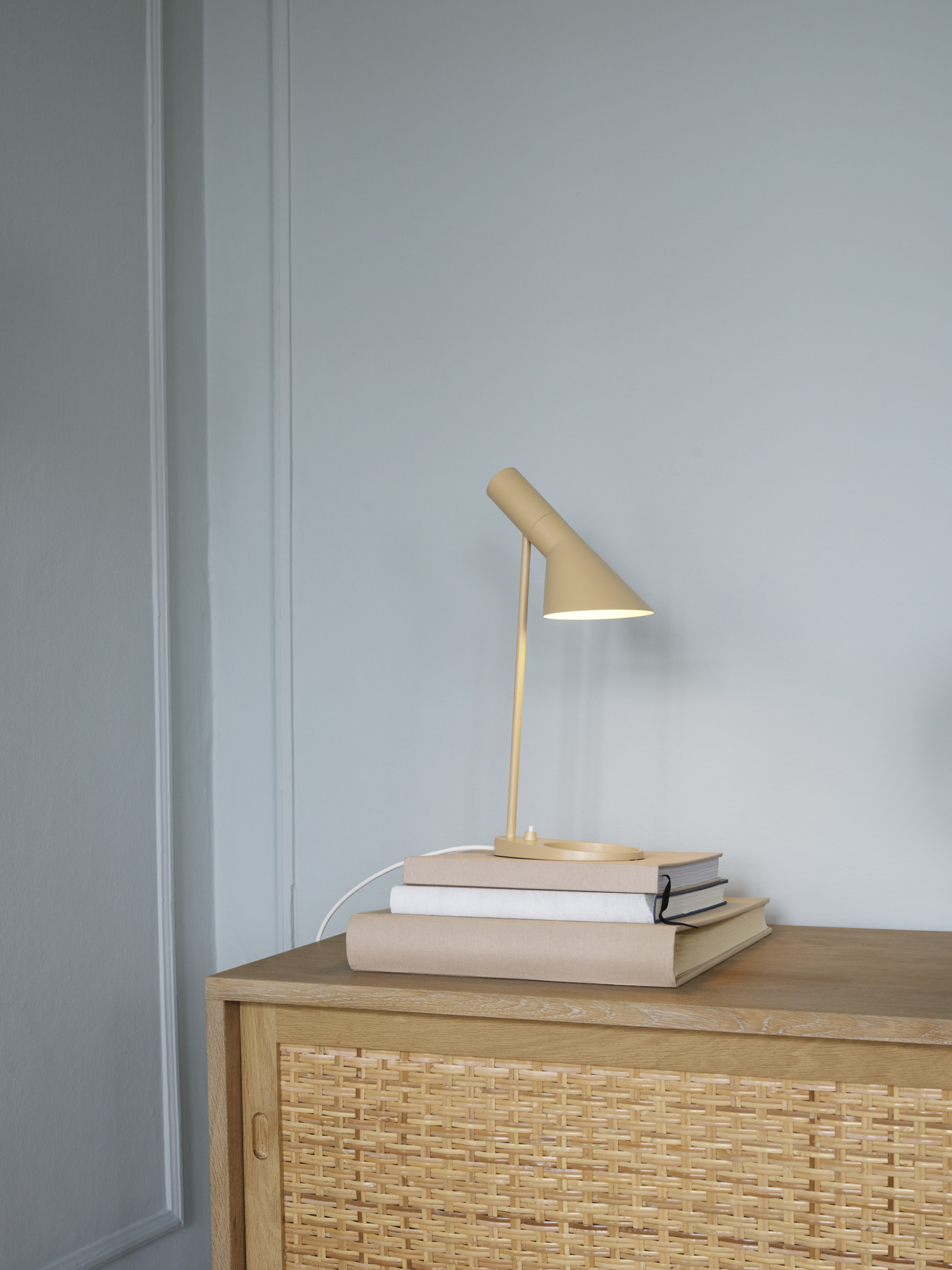 AJ Mini Table lamp in warm sand atop books on a light wooden dresser, © 2025 Louis Poulsen.